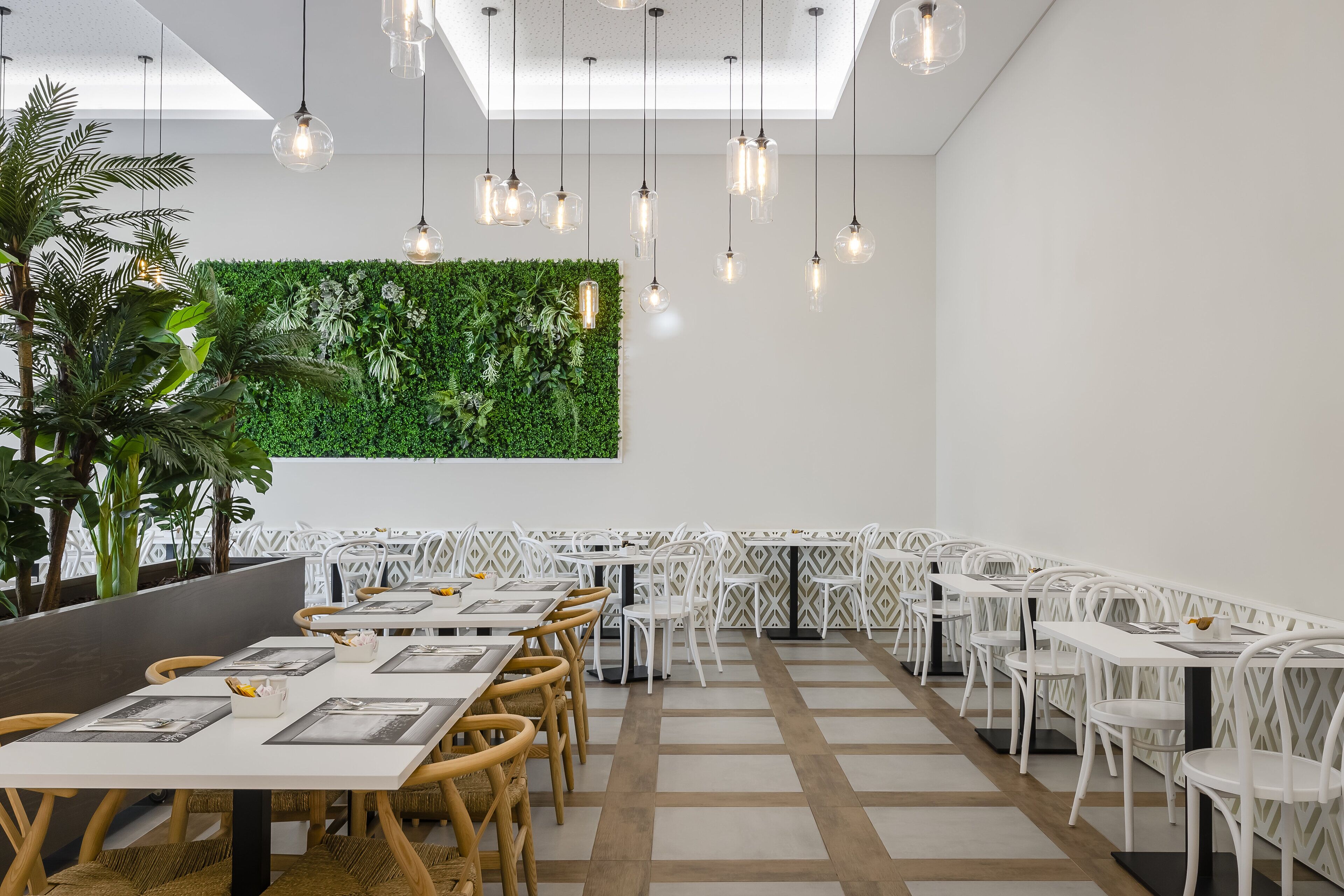 A modern dining area with white tables and chairs, a green wall with plants, and hanging pendant lights.