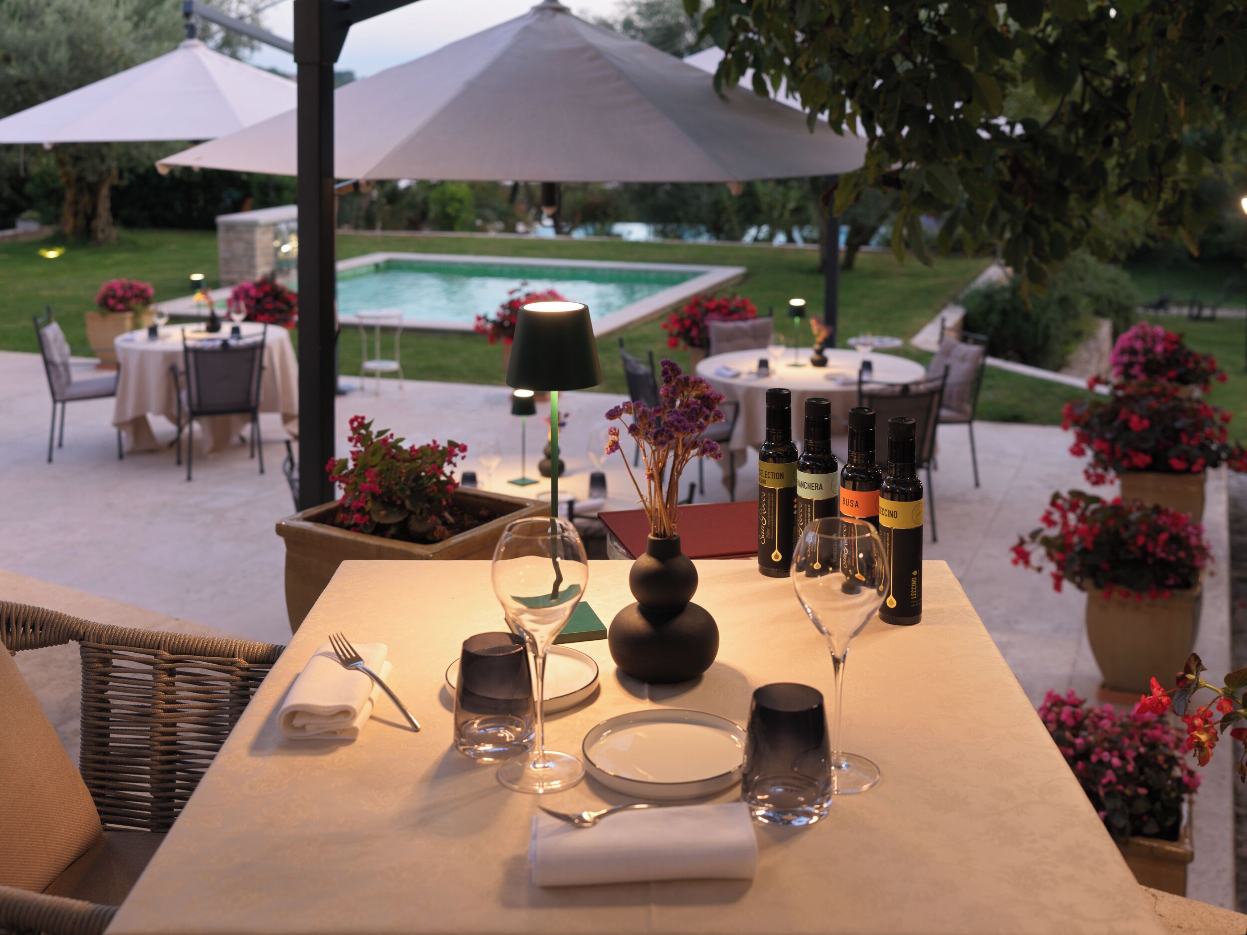 An outdoor dining area with a table set for two, a pool in the background, and umbrellas providing shade.
