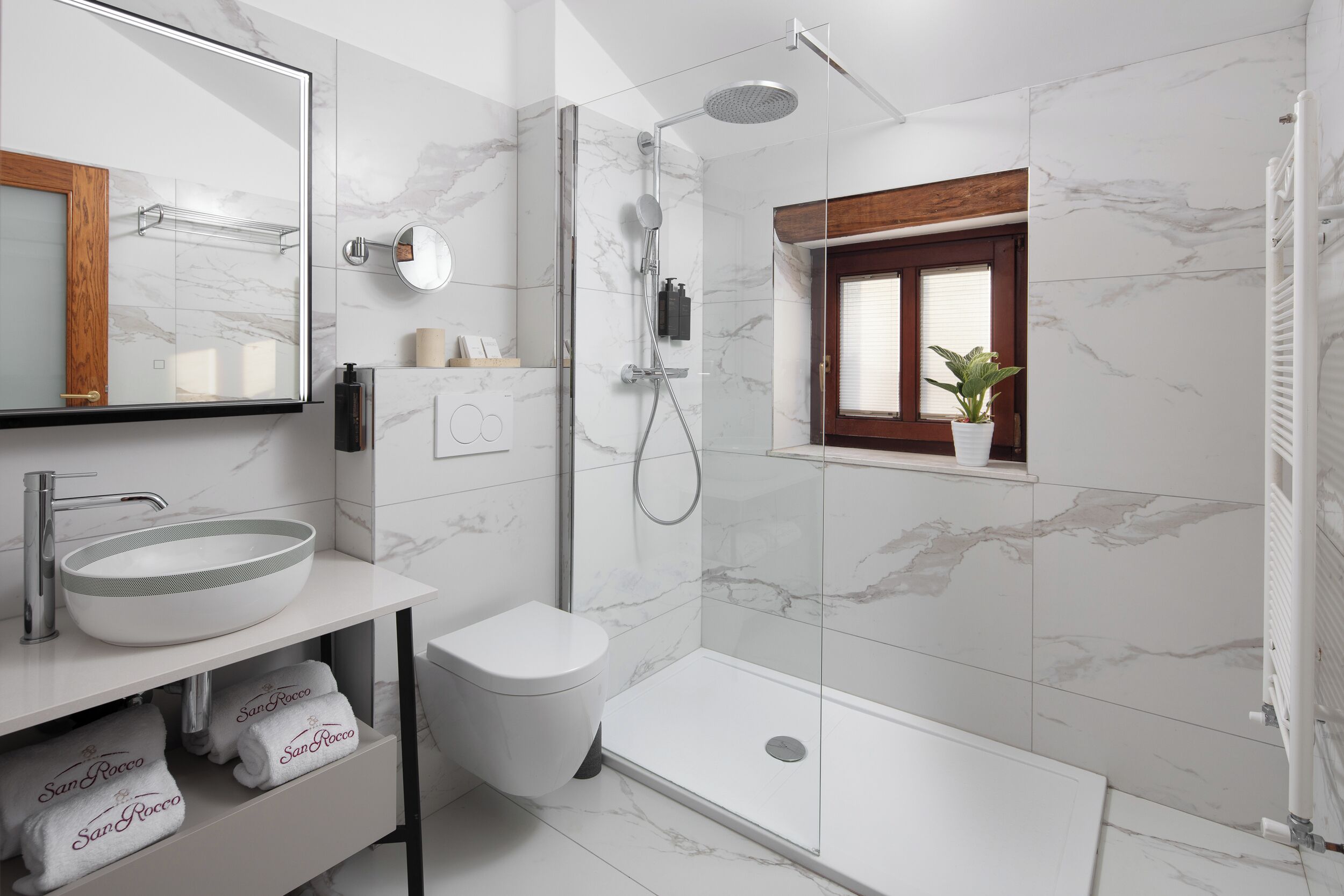 A modern bathroom with a large mirror, a round sink, a toilet, a shower with a glass door, and a small window with a plant.