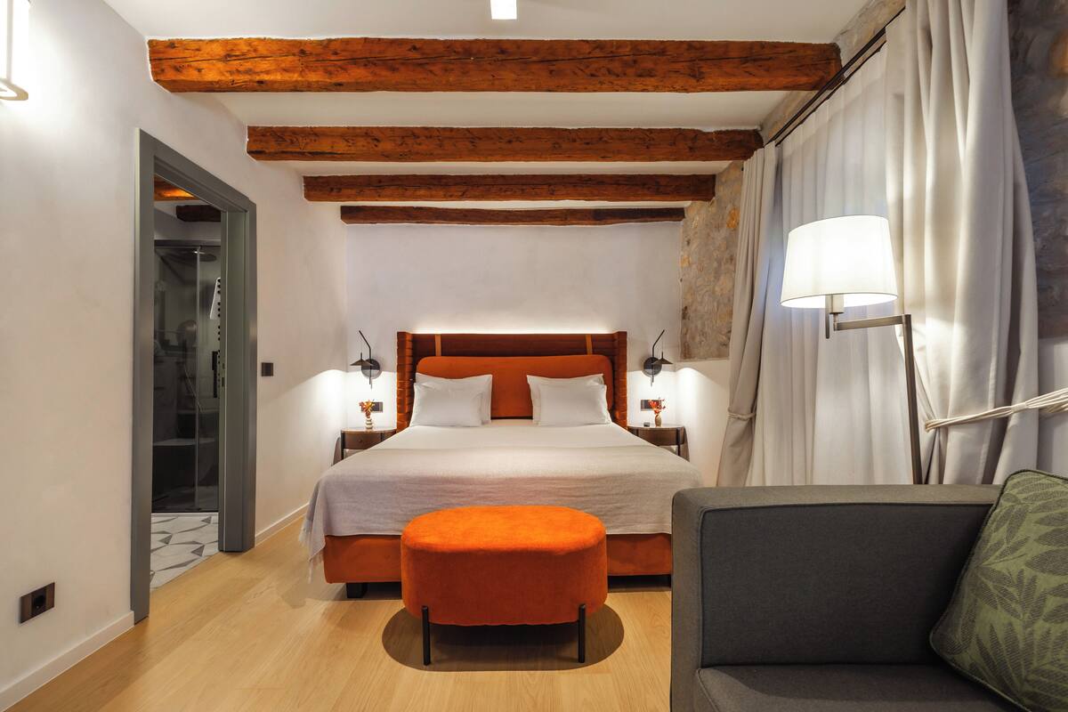 A bedroom with a wooden ceiling, a bed with white bedding, a nightstand with a lamp, and a sofa with a cushion.