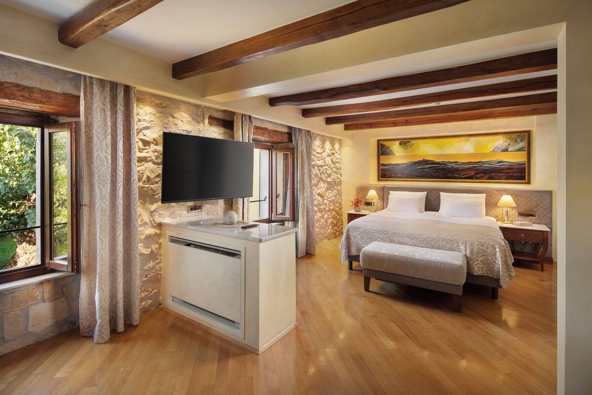 A bedroom with a bed, bedside tables, a TV, a window with curtains, and a painting on the wall.