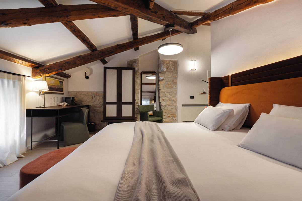 A hotel room with a bed, a desk, a chair, and a stone wall.