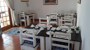 Un comedor con sillas blancas y una mesa negra dispuesta para cuatro. Hay dos cuadros en la pared, una ventana con cortinas y un piso de baldosas.