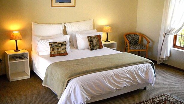 Una cama bien hecha con ropa blanca y almohadas decorativas, dos lámparas de noche, una silla de madera y un cuadro en la pared.