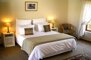 Una cama bien hecha con ropa blanca y almohadas decorativas, dos lámparas de noche, una silla de madera y un cuadro en la pared.