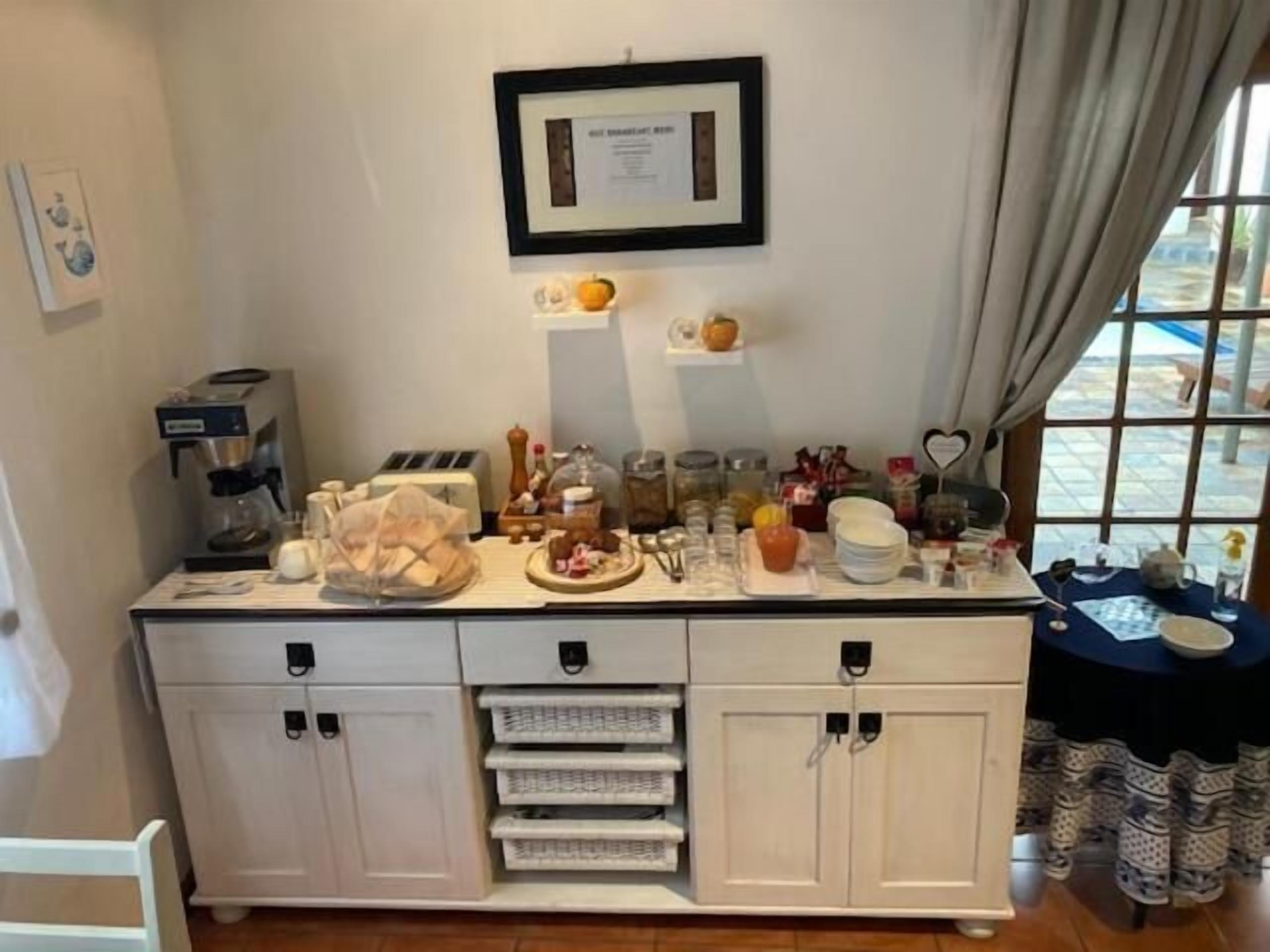 A kitchen area with a coffee machine, toaster, and various food items on the counter. There are cabinets below the counter and a window with curtains on the right side.
