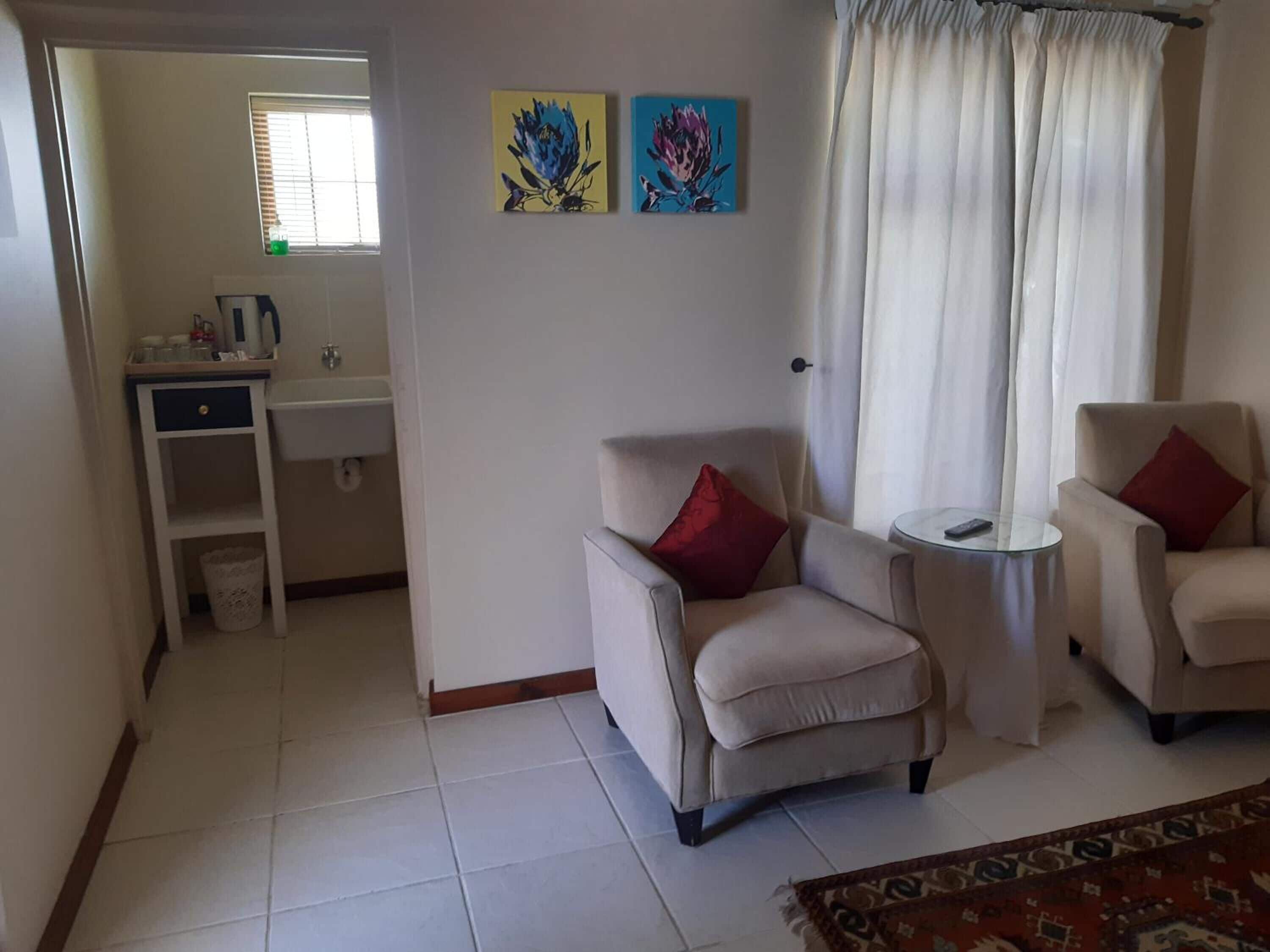 A living room with two armchairs, a small round table, and a sofa. There is a window with blinds, a sink, and a trash bin in the adjacent room. Two paintings are hung on the wall.