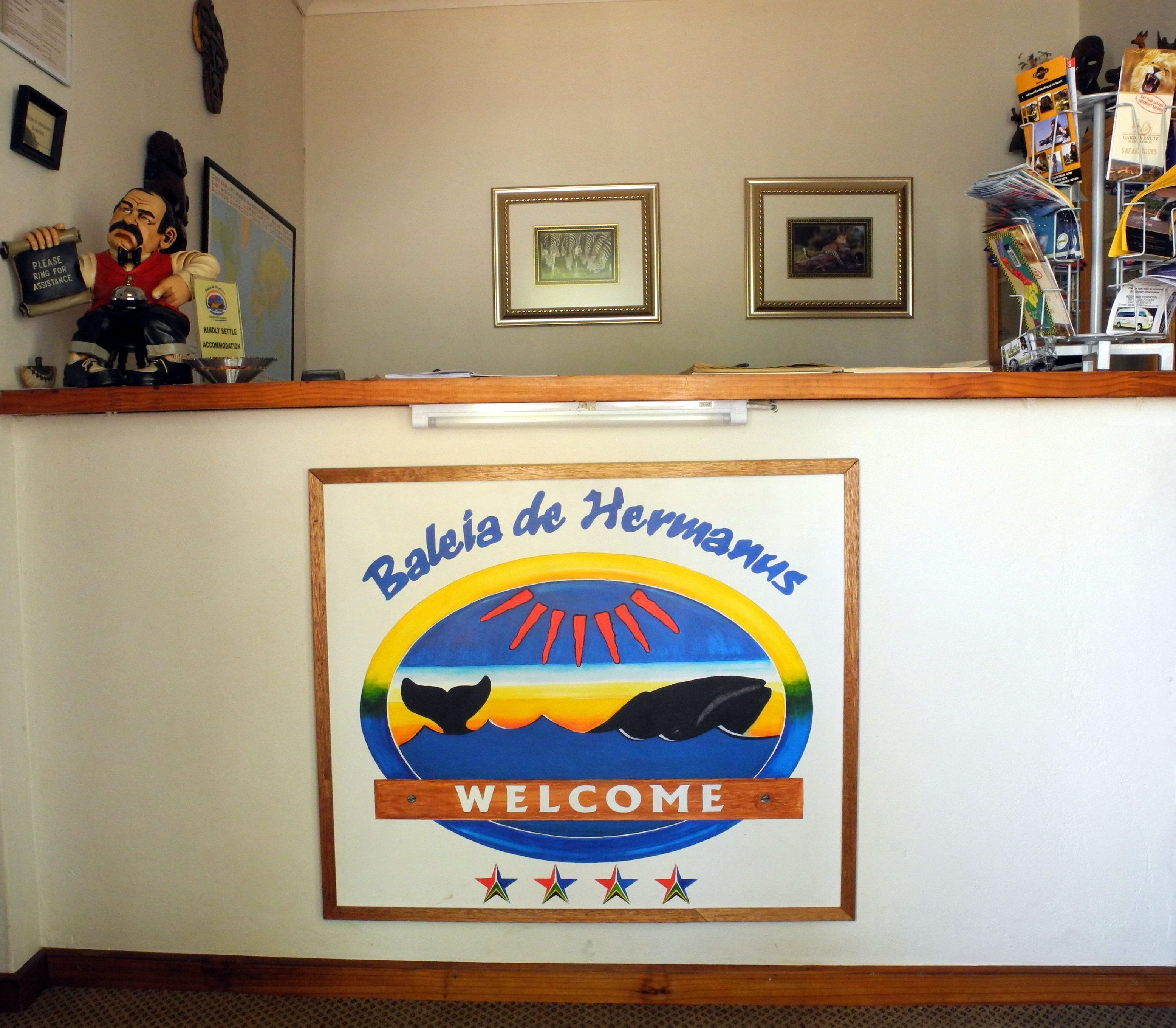 Un comptoir d’accueil avec une pancarte de bienvenue pour Baleia de Hermanus, représentant une baleine et un coucher de soleil.