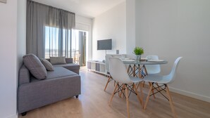 Un salon moderne avec un canapé gris, une table à manger entourée de chaises blanches, un téléviseur à écran plat fixé au mur et une grande fenêtre agrémentée de rideaux.