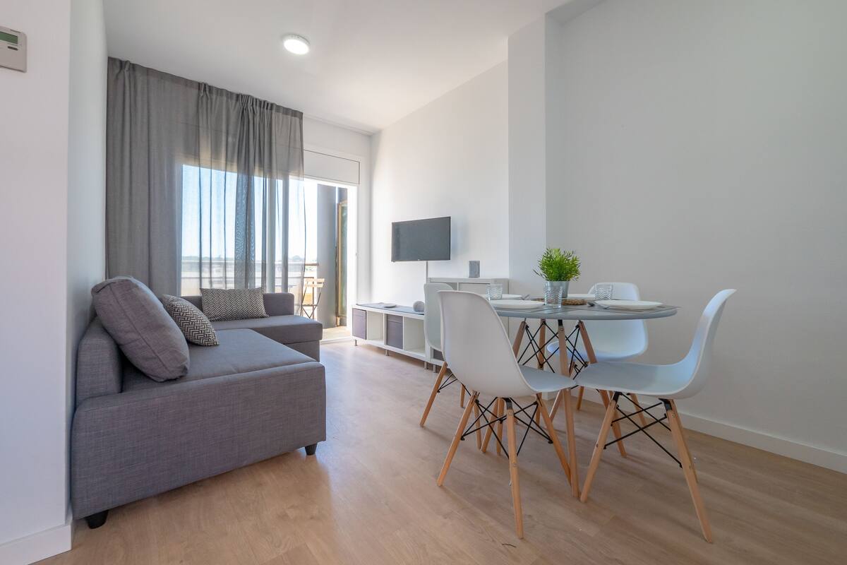 Ein modernes Wohnzimmer mit grauem Sofa, einem Esstisch mit weißen Stühlen, einem flachbildfernseher an der Wand und einem großen Fenster mit Vorhängen.