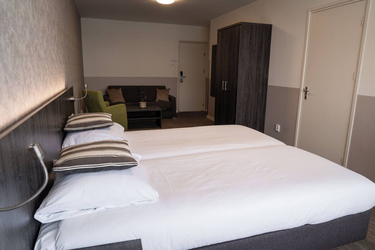 A neatly made bed with white bedding and striped pillows, a green armchair, and a dark wooden wardrobe in a hotel room.