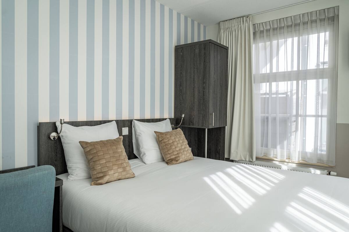 A hotel room with a bed, two pillows, a nightstand, a chair, a wardrobe, and a window with curtains.