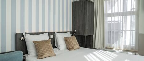 A hotel room with a bed, two pillows, a nightstand, a chair, a wardrobe, and a window with curtains.