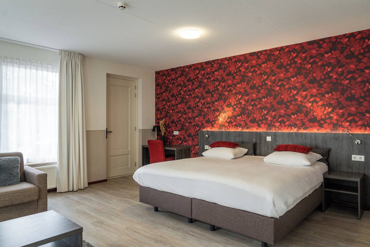 A modern hotel room with a large bed, a desk, a chair, and a red floral wallpaper.