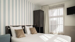 A neatly made bed with white linens and two pillows, a dark wooden wardrobe, a television mounted on the wall, and a window with sheer curtains allowing natural light. - Het Hart van Weesp (Weesp)