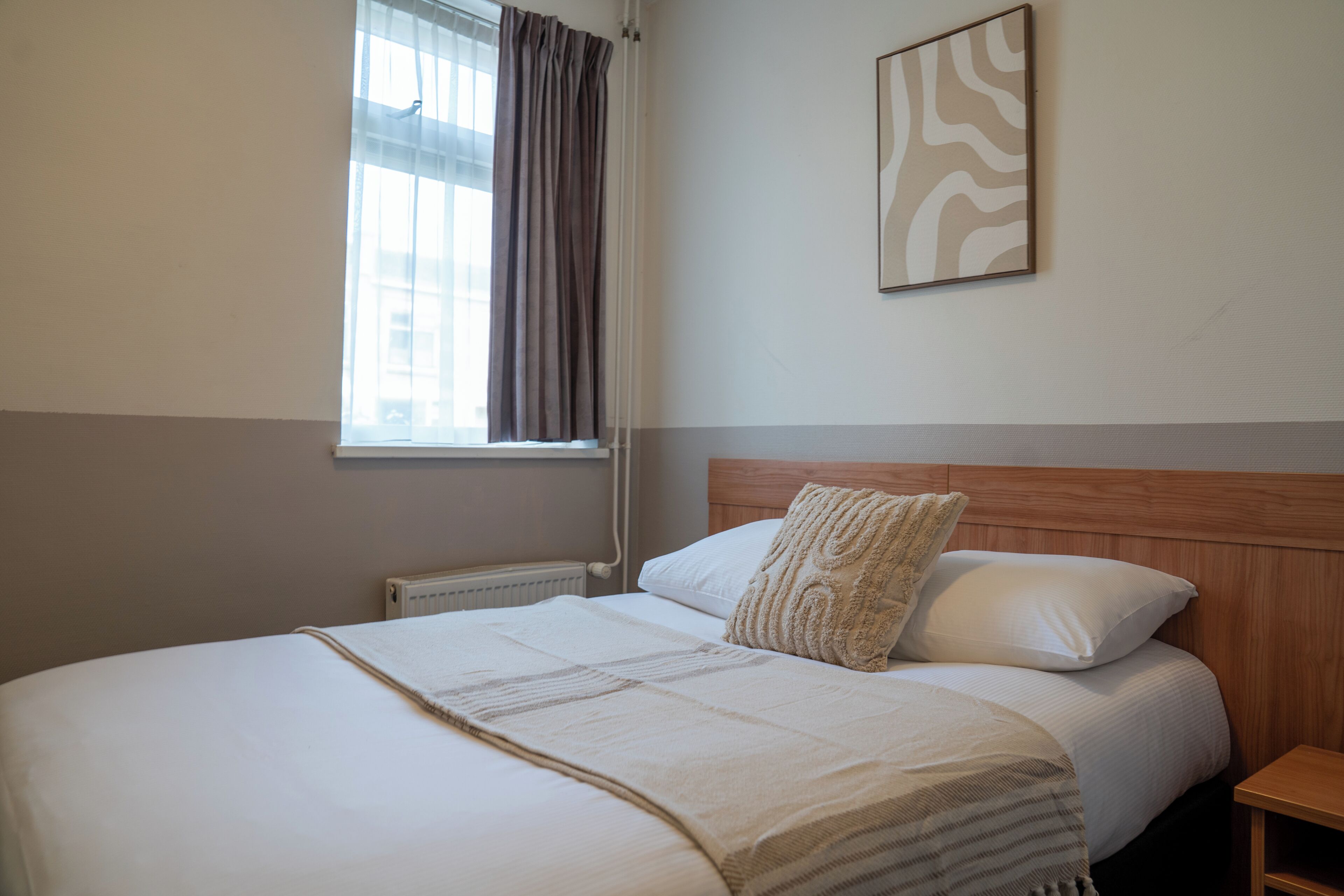 A neatly made bed with a headboard, a bedside table, a window with curtains, and a wall-mounted artwork.