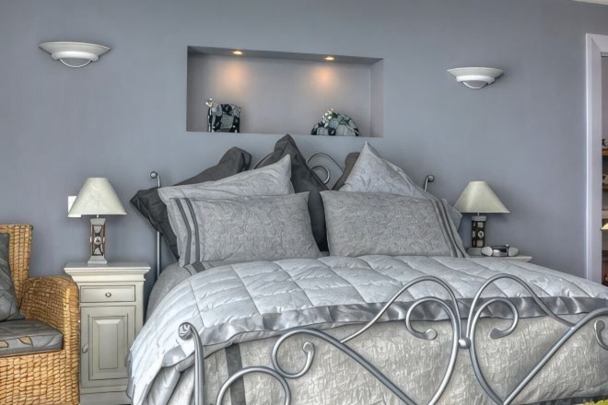 A bedroom with a metal bed frame, grey bedding, two bedside lamps, a wicker chair, and a small bedside table.