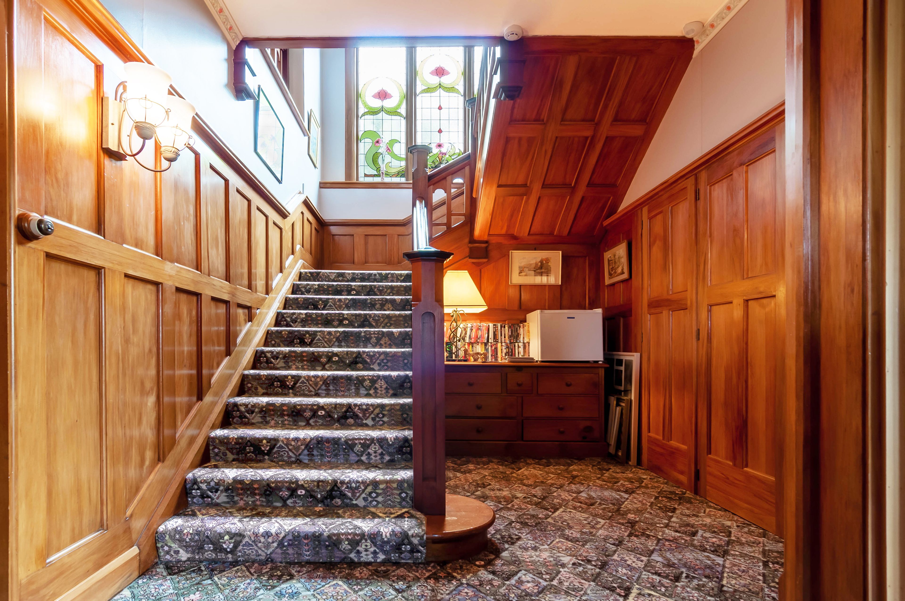 A wooden staircase with patterned carpet, a wooden handrail, and a view of a room with a refrigerator and a window with stained glass.