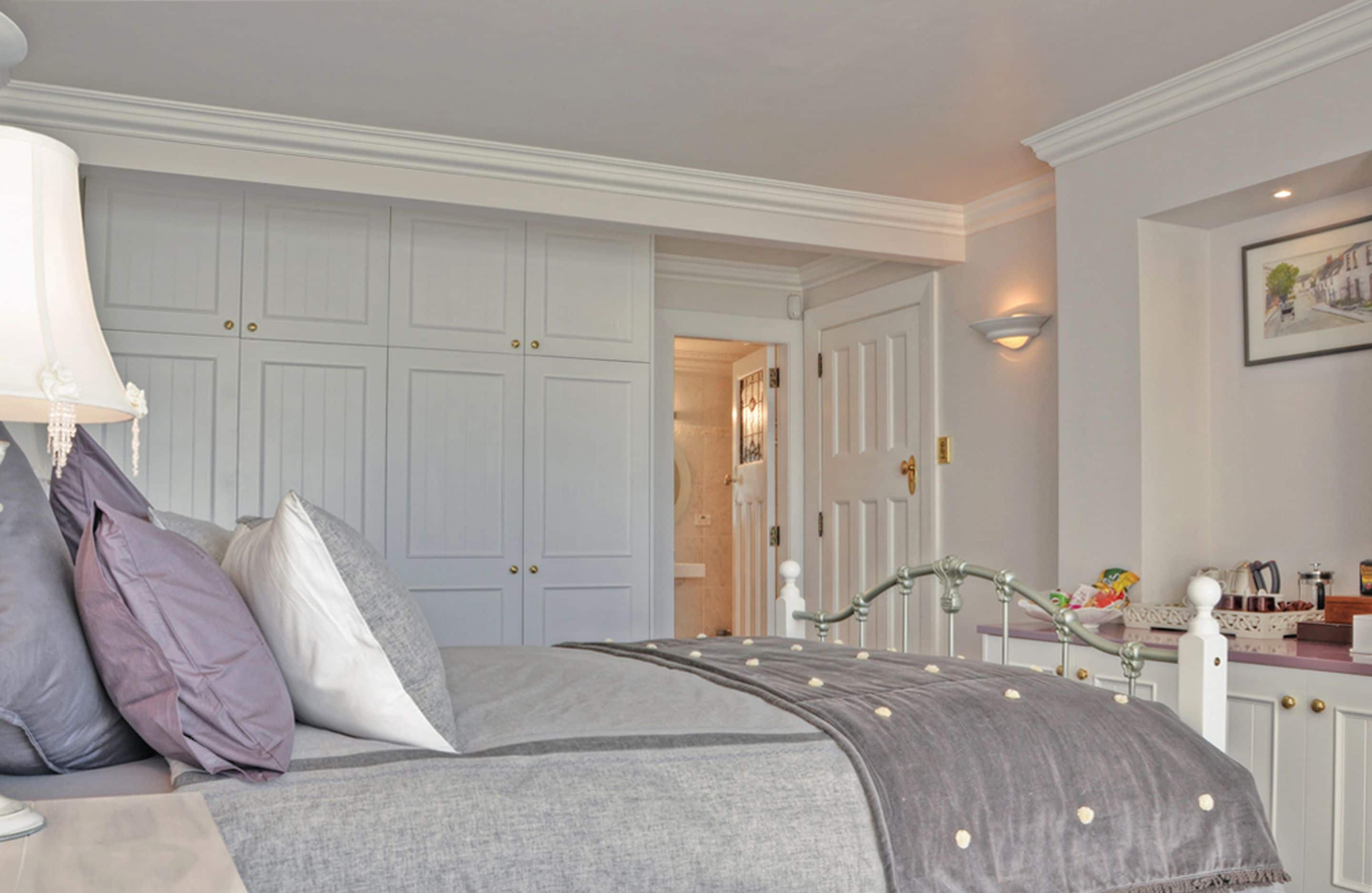Un dormitorio con cama, mesita de noche, lámpara y cómoda. Se ve una puerta que da a otra habitación.