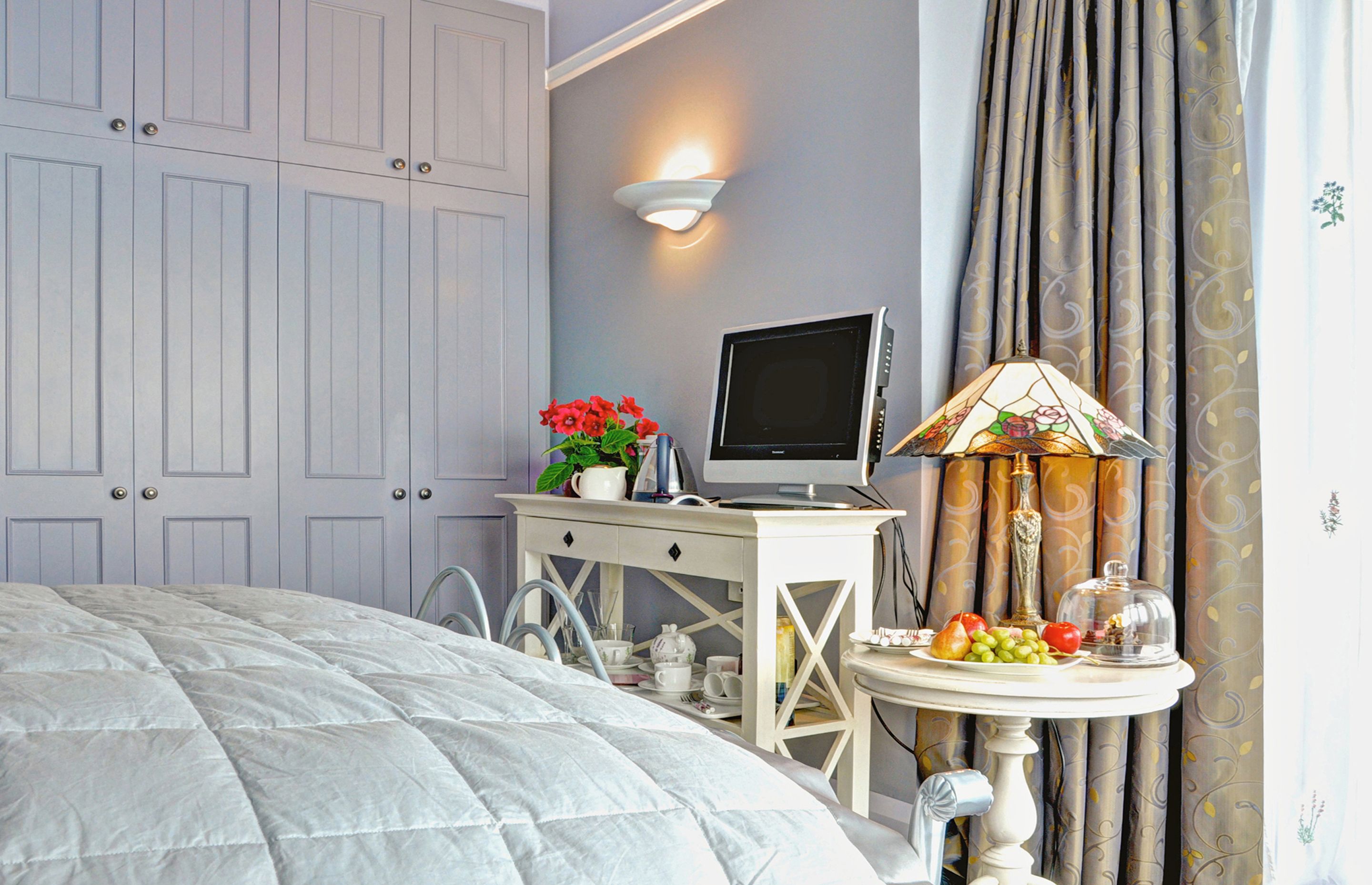 A bedroom with a bed, a desk with a computer, a lamp, and a table with fruits and a vase of flowers.