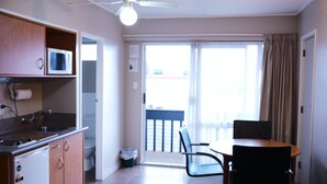 A compact kitchen with a microwave, sink, and a ceiling fan. A dining area with a table and chairs is visible, and a balcony with a view is in the background. - Kiwi Studios Motel (Palmerston North)