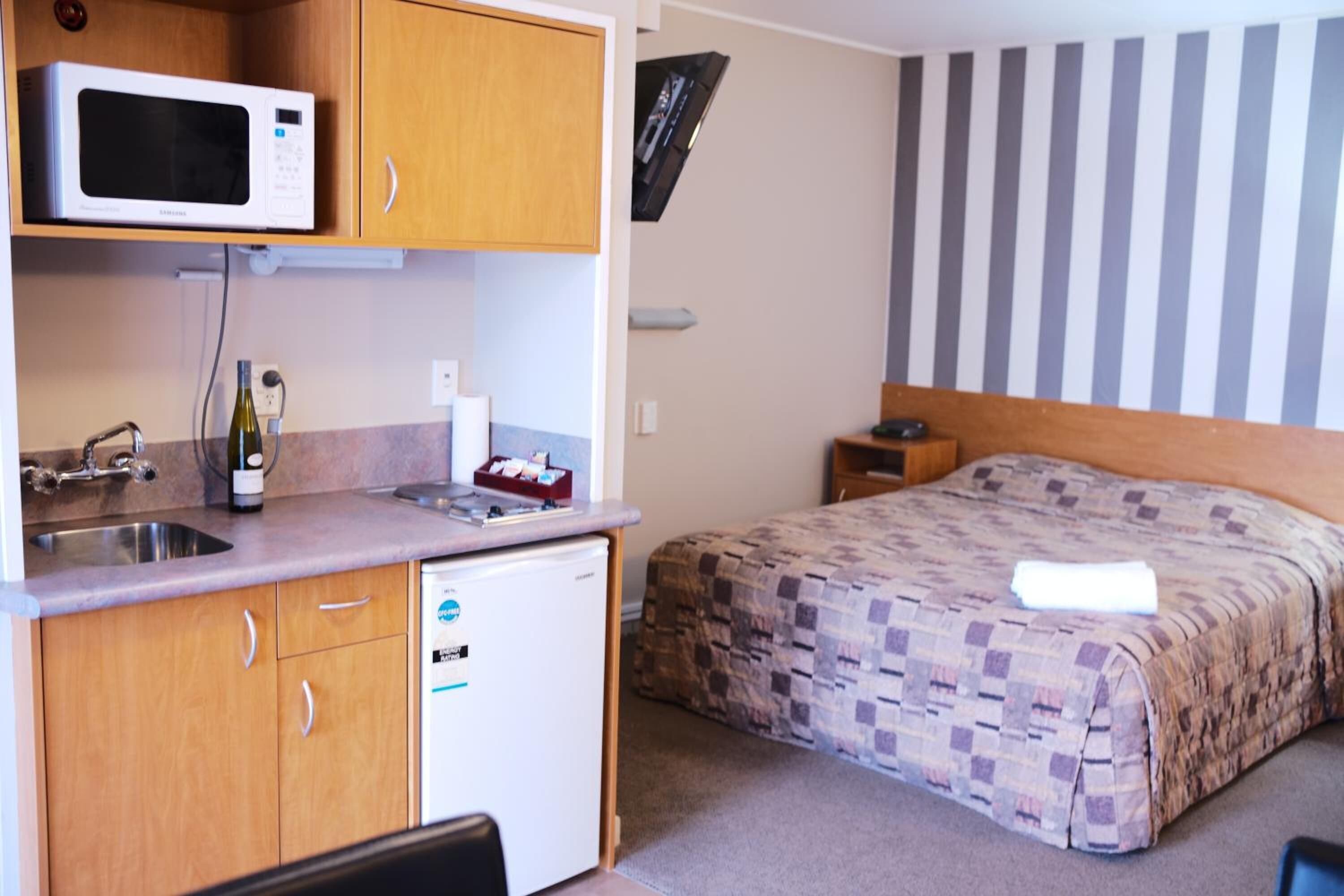 A compact hotel room with a kitchenette, a bed with a patterned bedspread, and a striped wall.