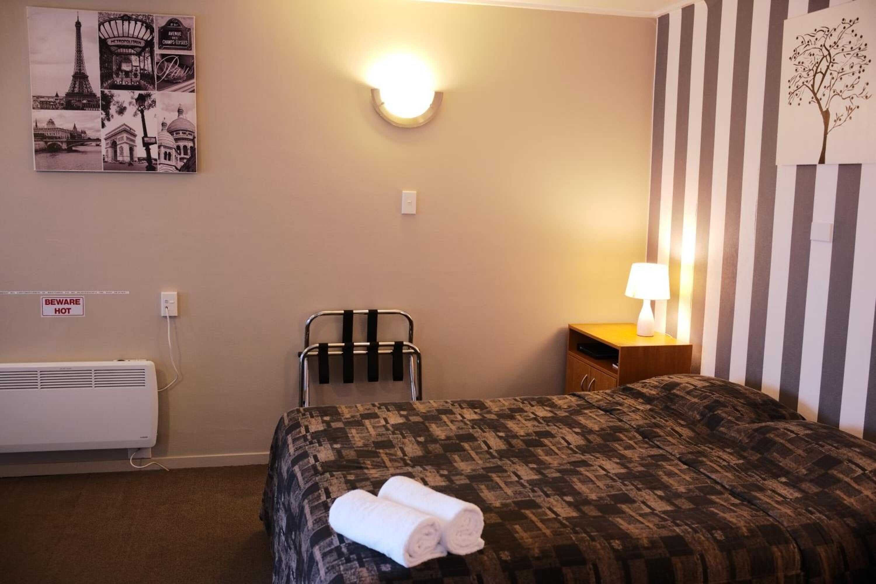 Ett hotellrum med en säng, en stol, ett nattduksbord, en lampa, en väggmonterad lampa och en affisch med bilder av kända landmärken i Paris.
