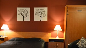A hotel room with a bed, two bedside tables with lamps, a wardrobe, and a chair with a cushion. - Kiwi Studios Motel (Palmerston North)