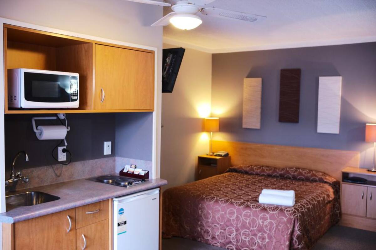 A compact hotel room with a kitchenette, a bed with a patterned bedspread, bedside lamps, and a ceiling fan.