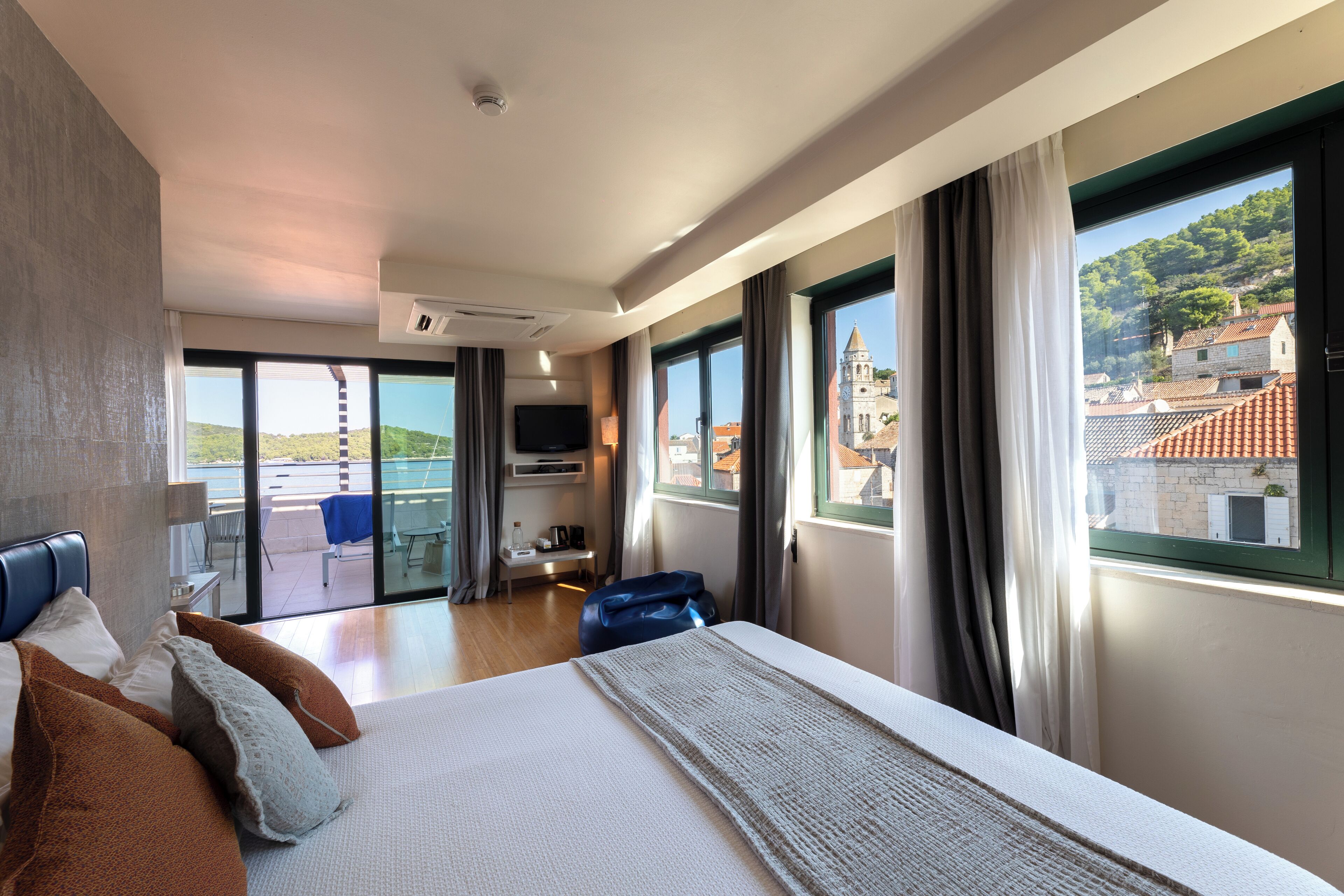 A hotel room with a bed, a desk, a chair, and a view of a town with buildings and a hill.