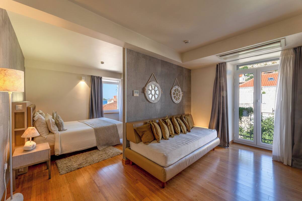 Un dormitorio moderno con cama, sofá, mesita de noche y un amplio ventanal con cortinas.