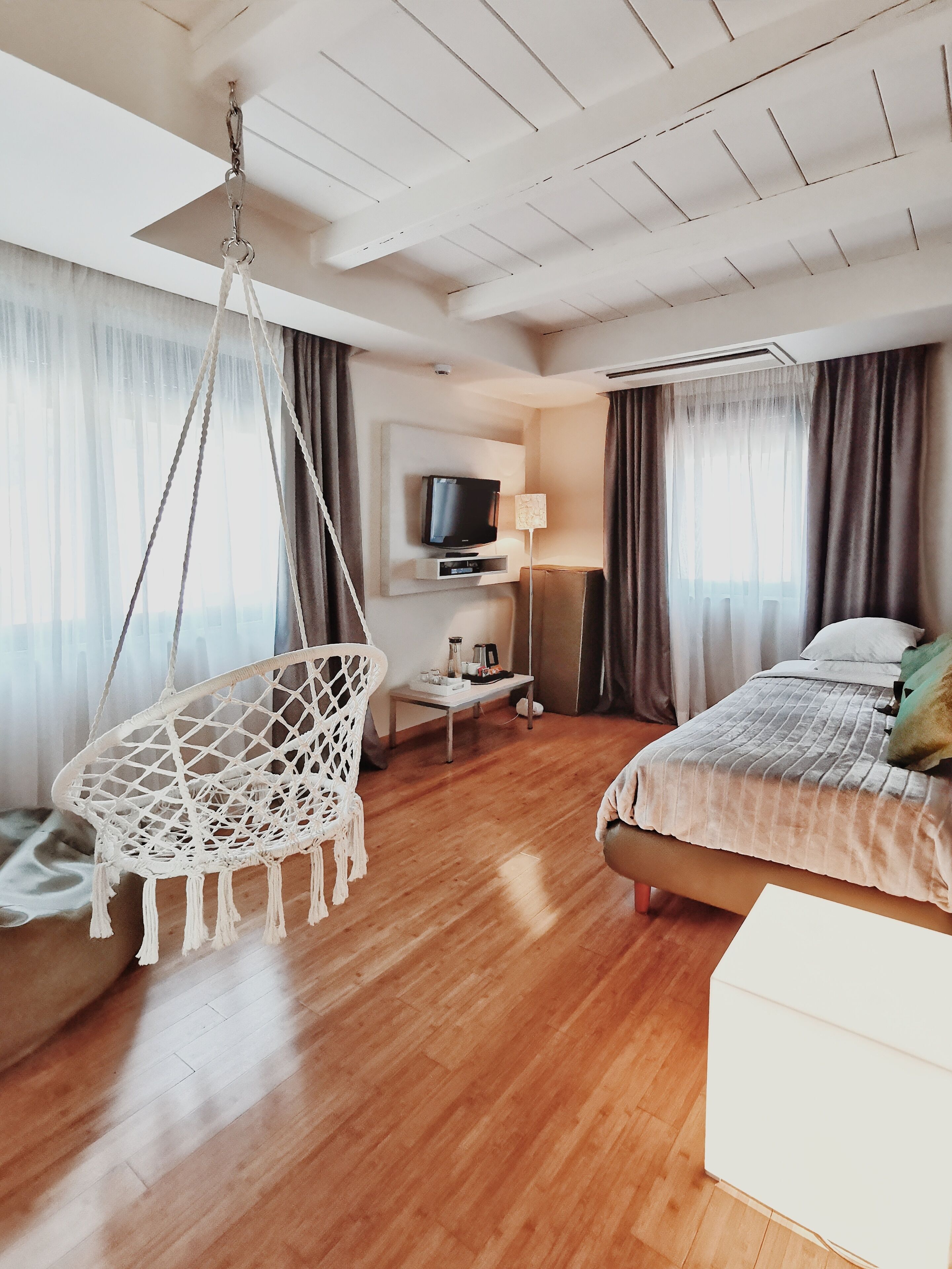 A hotel room with a wooden floor, a hanging chair, a bed with a white headboard, a television mounted on the wall, and a window with curtains.