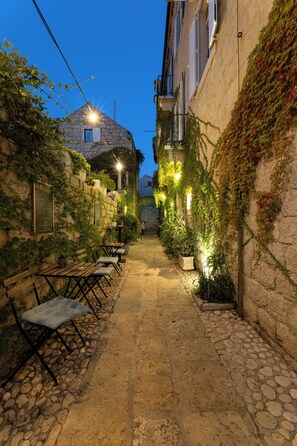 Une ruelle étroite, pavée de pierres, bordée de bancs et agrémentée de plantes en pot, avec des bâtiments aux murs recouverts de lierre et dotés d’un éclairage extérieur.