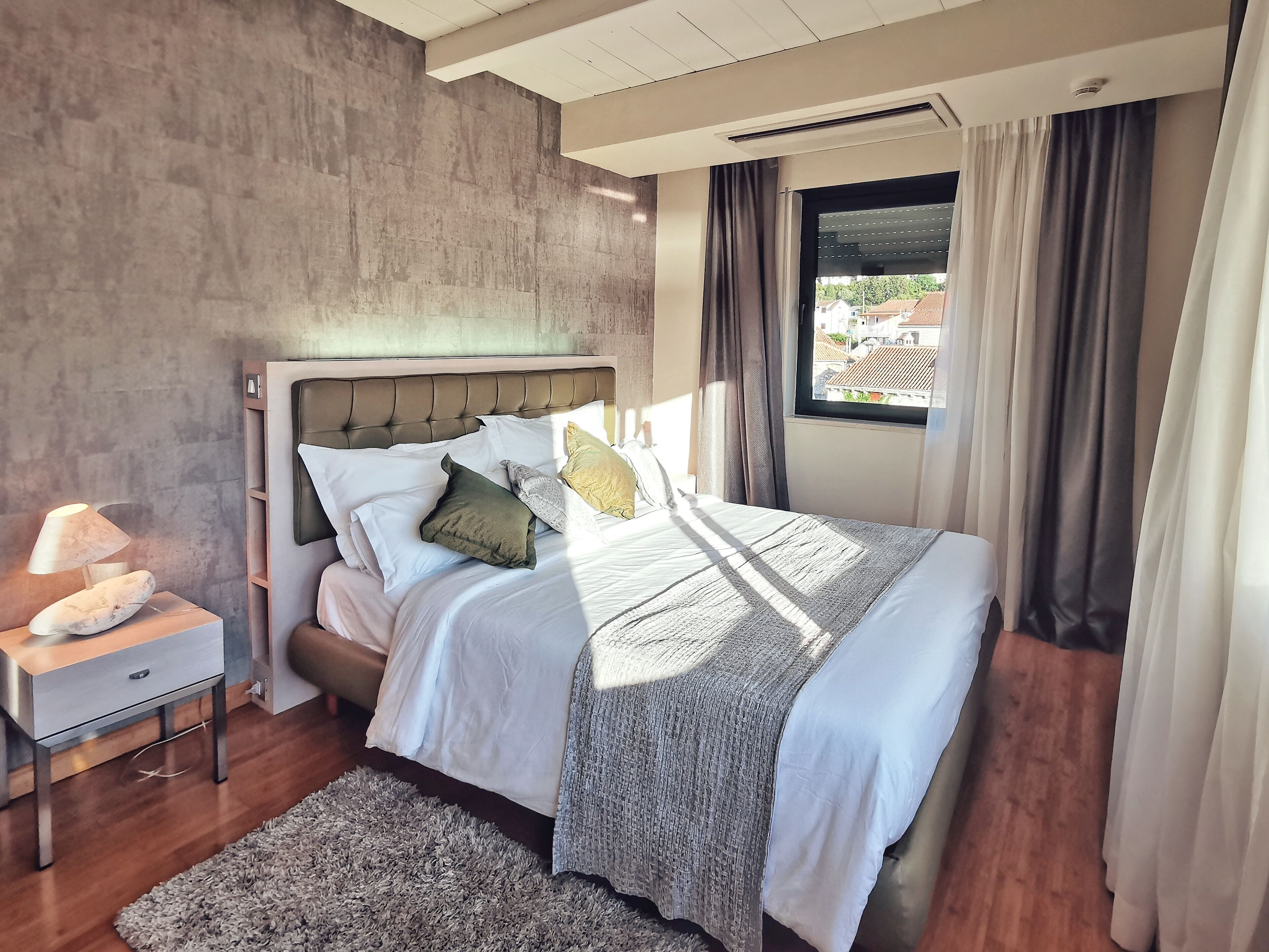 Ein modernes Schlafzimmer mit einem großen Bett, einem Nachttisch mit Lampe und Blick ins Freie durch ein Fenster mit Vorhängen.