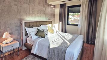 A modern bedroom with a large bed, a bedside table with a lamp, and a view of the outside through a window with curtains.