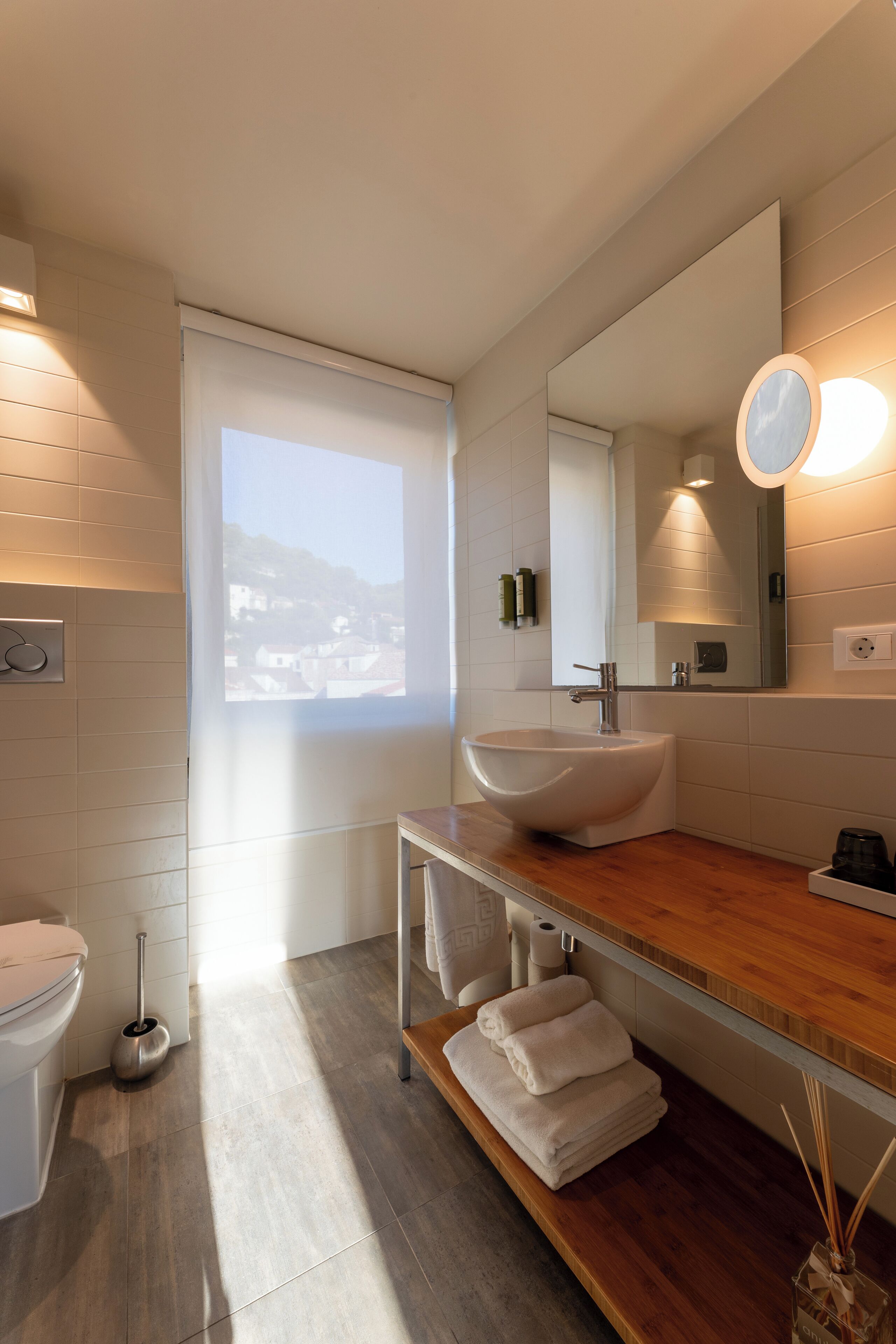 A modern bathroom with a large window, a white sink, and a wooden vanity.