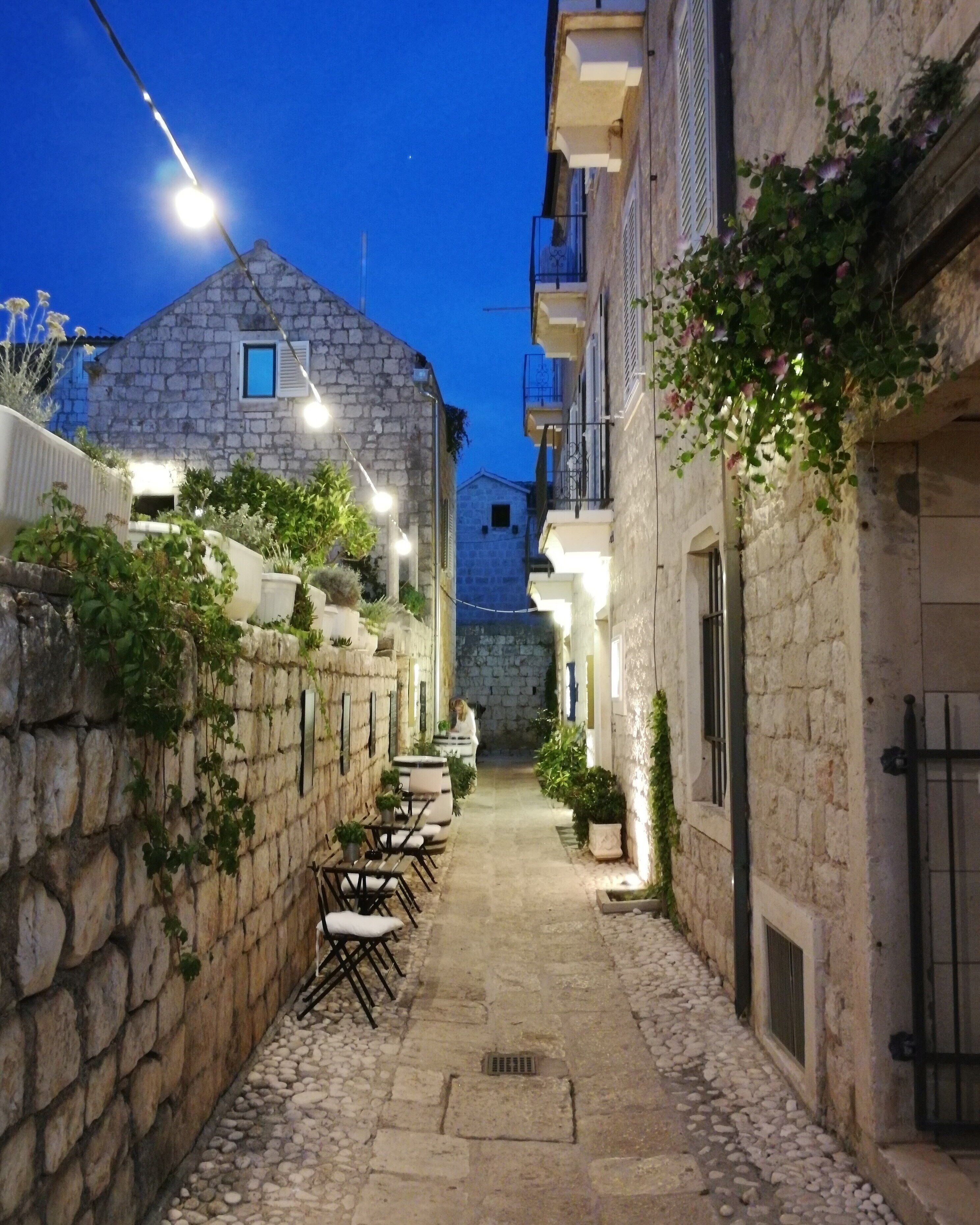 Une ruelle étroite pavée, avec des tables à l’extérieur et des guirlandes lumineuses, qui mène à un bâtiment en pierre doté d’un toit en pente.