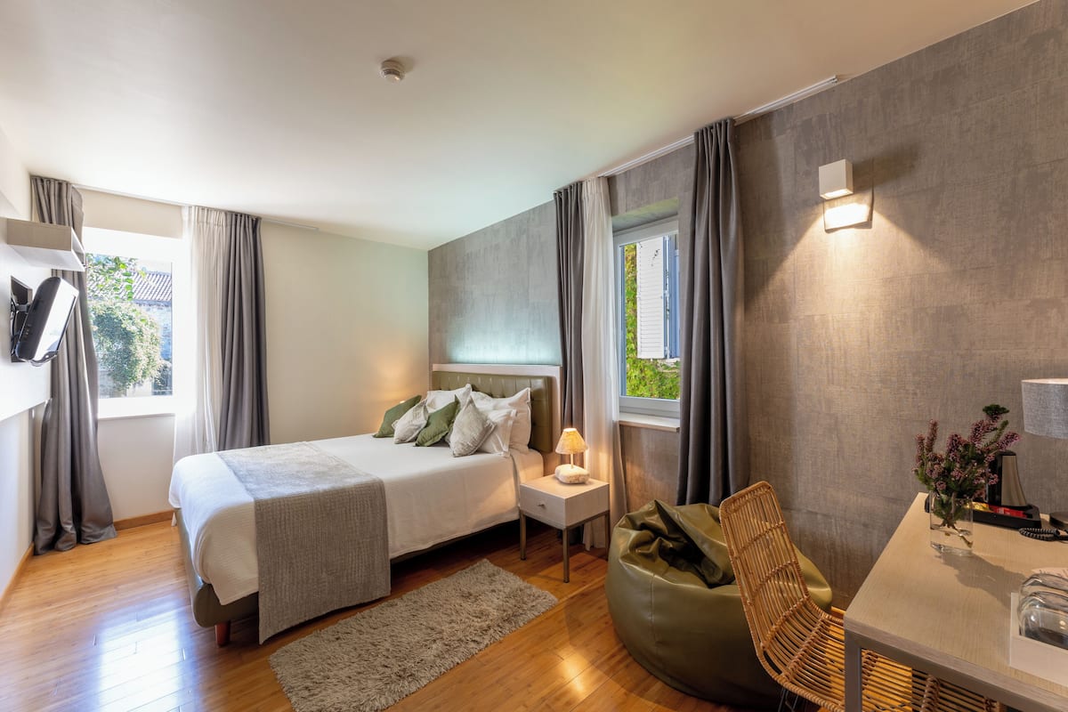 Un dormitorio moderno con cama, mesita de noche, lámpara, silla y vistas al verdor exterior.