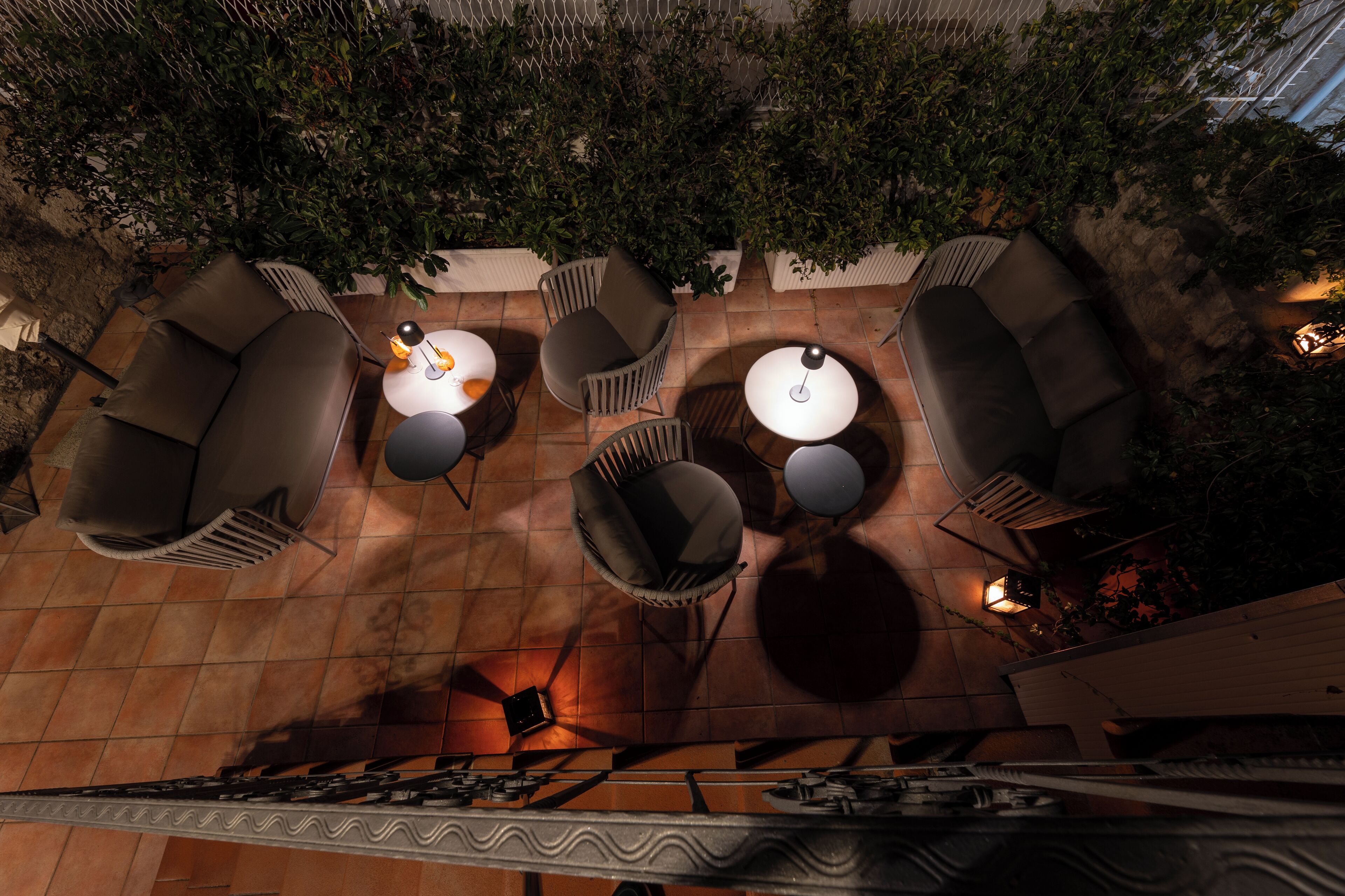 An outdoor patio with multiple seating areas, including sofas and round tables, surrounded by greenery and stone flooring.