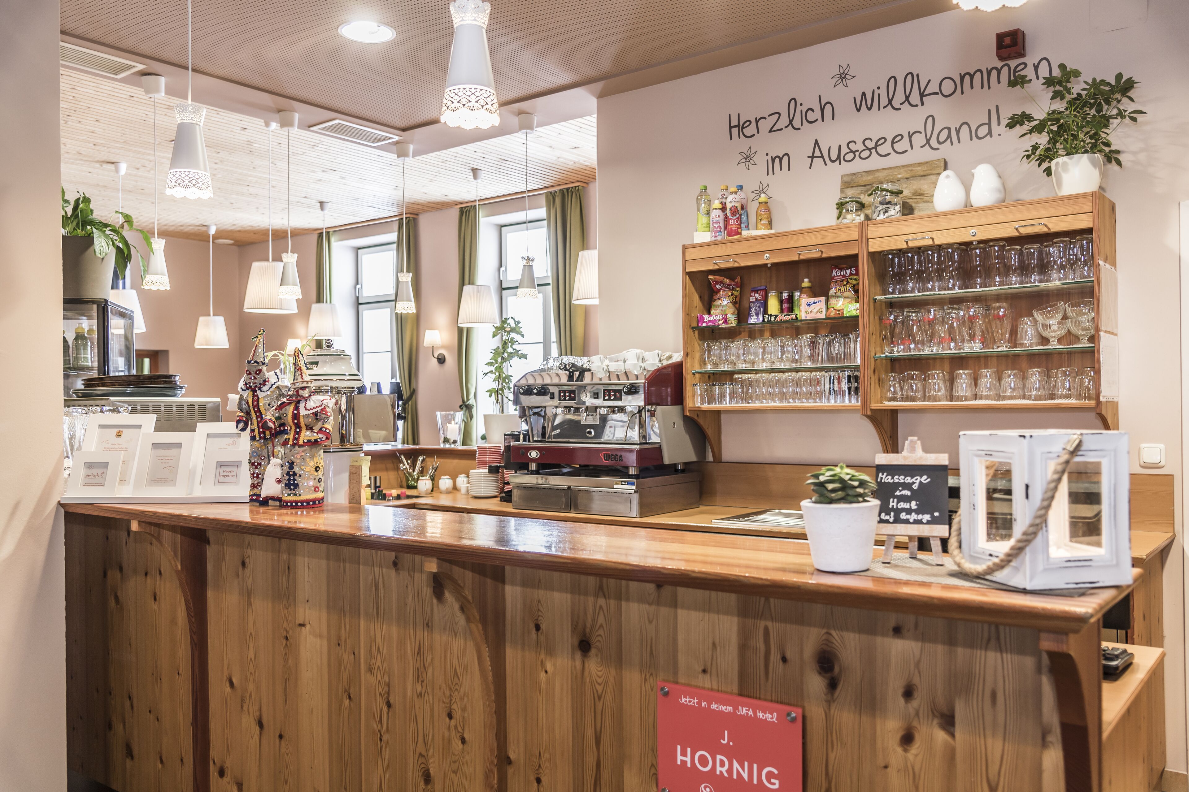 Zona de recepción de hotel con mostrador de madera, una máquina de café y un cartel de bienvenida a los huéspedes.