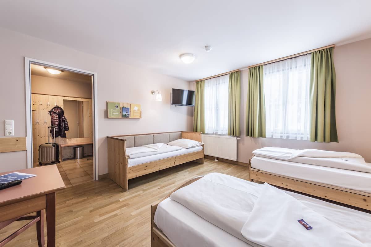 Una habitación de hotel con dos camas individuales, un escritorio de madera, un televisor y un baño visible a través de una puerta abierta.