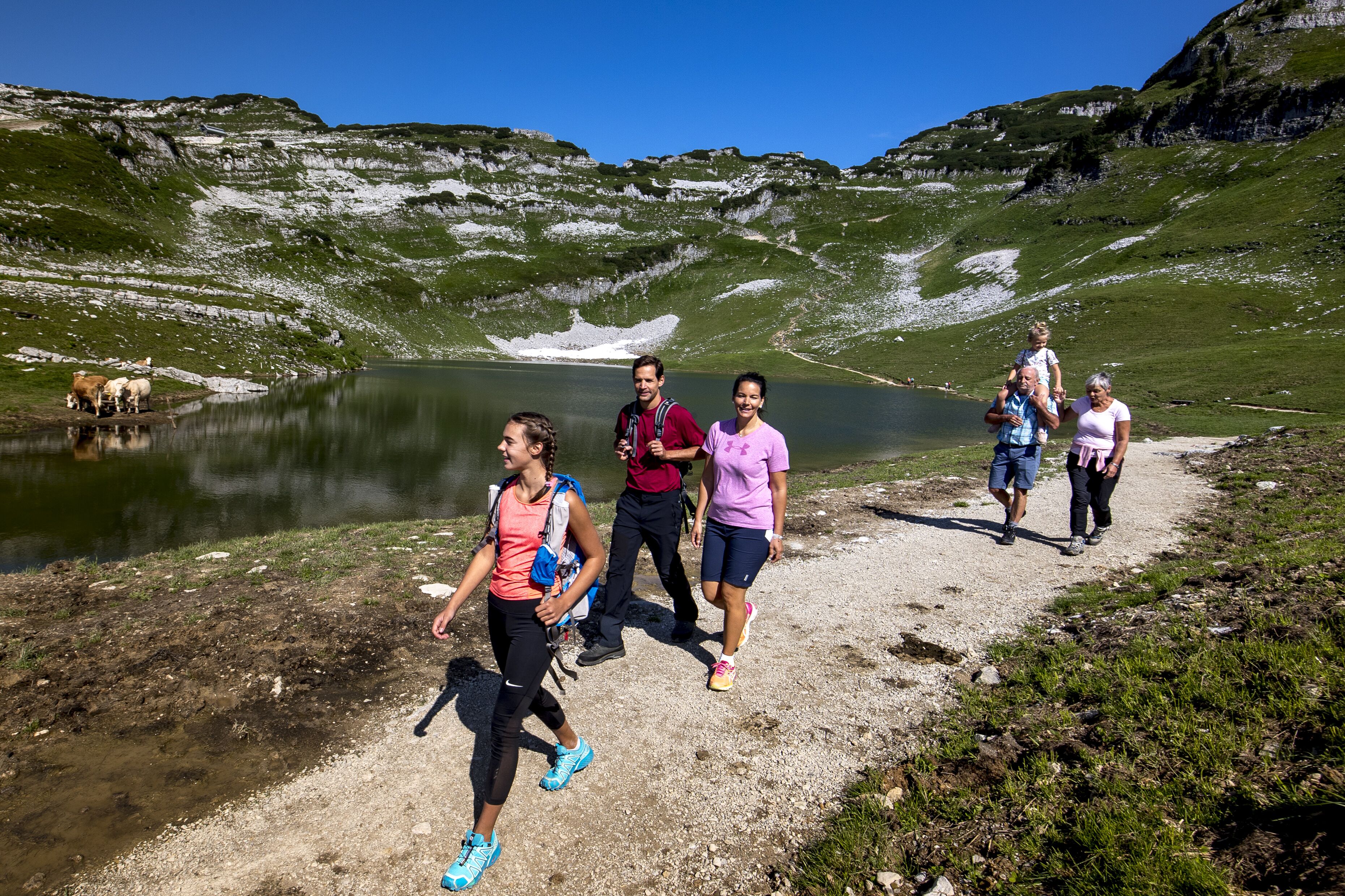 Un grupo de personas caminando por un sendero junto a un lago, con montañas al fondo.