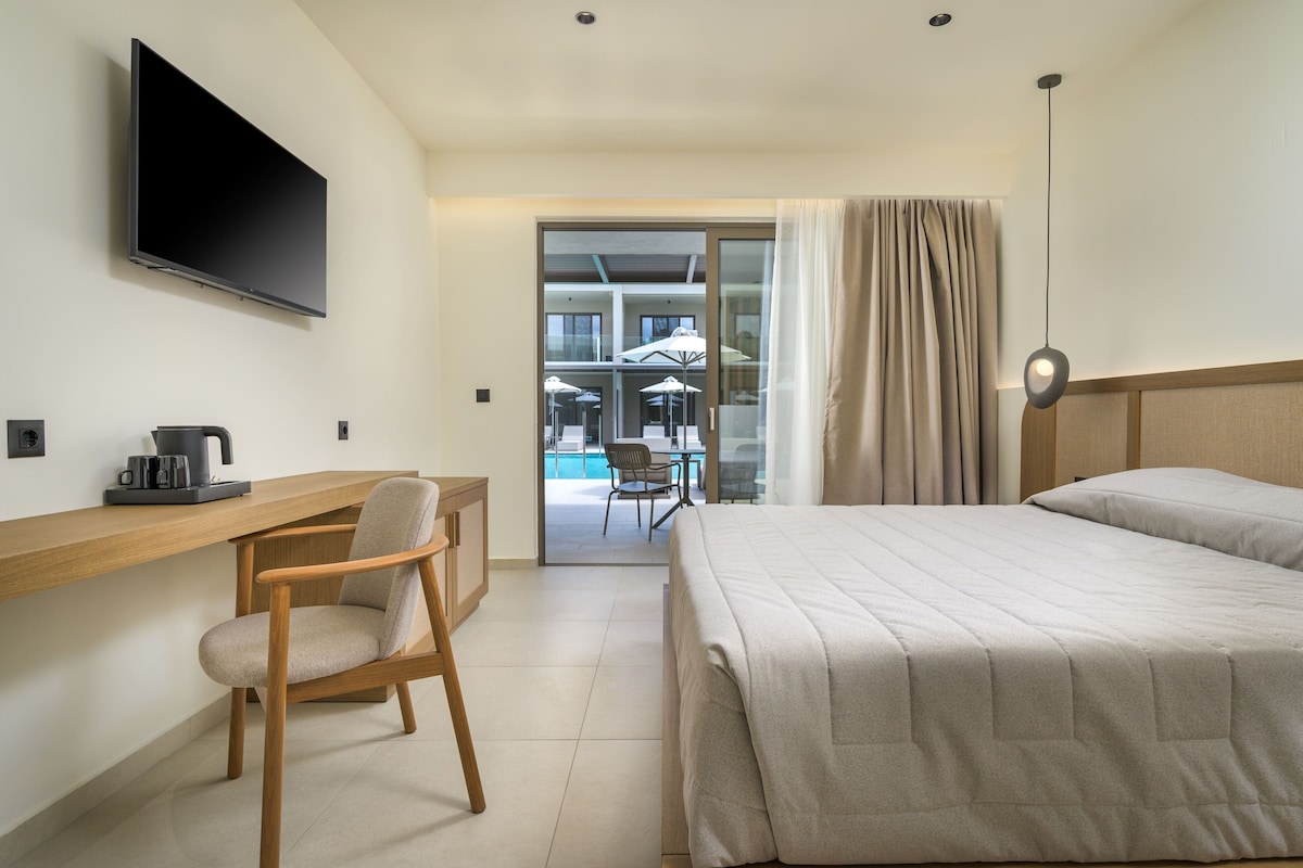 A hotel room with a bed, a desk with a chair, a TV, and a view of an outdoor pool area.
