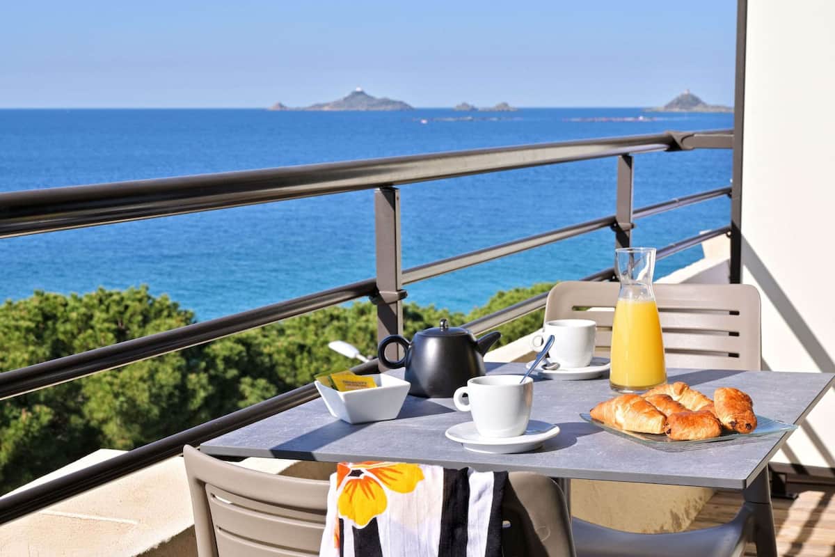 Un balcón con una mesa dispuesta para el desayuno, incluyendo croissants, una taza de café, una tetera y un vaso de zumo de naranja, con vistas al mar.