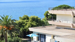 Ein Küstengebäude mit Balkon, der Blick auf ein klares blaues Meer und Hügel im Hintergrund bietet.