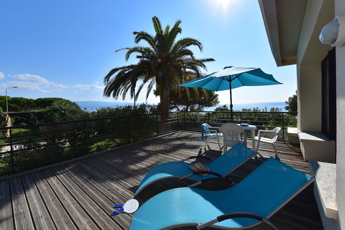 Una terraza de madera con sillones y una mesa, una palmera y vista al mar.