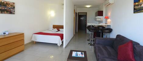 A hotel room with a bed, a wooden dresser, a brown wicker ottoman, a dark brown sofa with a red pillow, a kitchen area with a red lamp, and a dining area with a bar stool.