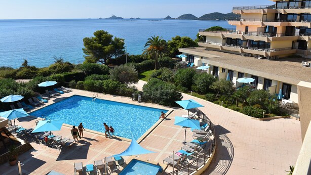 Ein Poolbereich mit mehreren Liegestühlen und Sonnenschirmen, freier Blick auf das Meer und ein Gebäude mit Balkonen, das auf den Pool blickt.