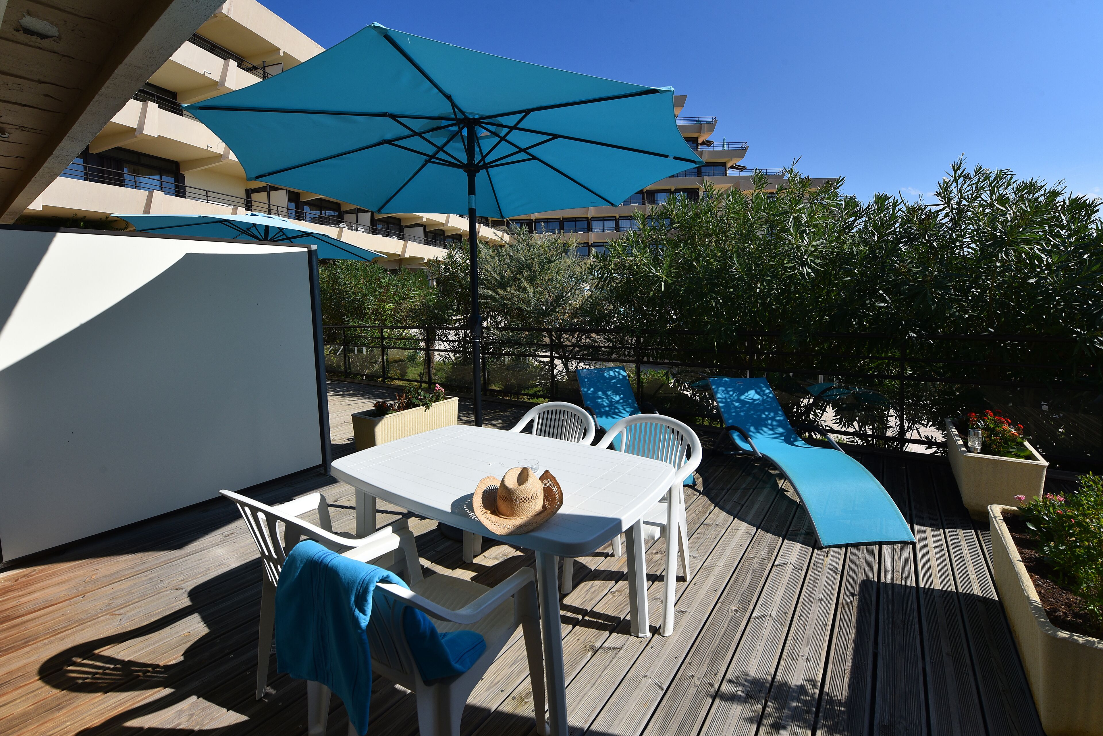 Una terraza de madera con una mesa, sillas, un paraguas azul y un sombrero sobre la mesa.