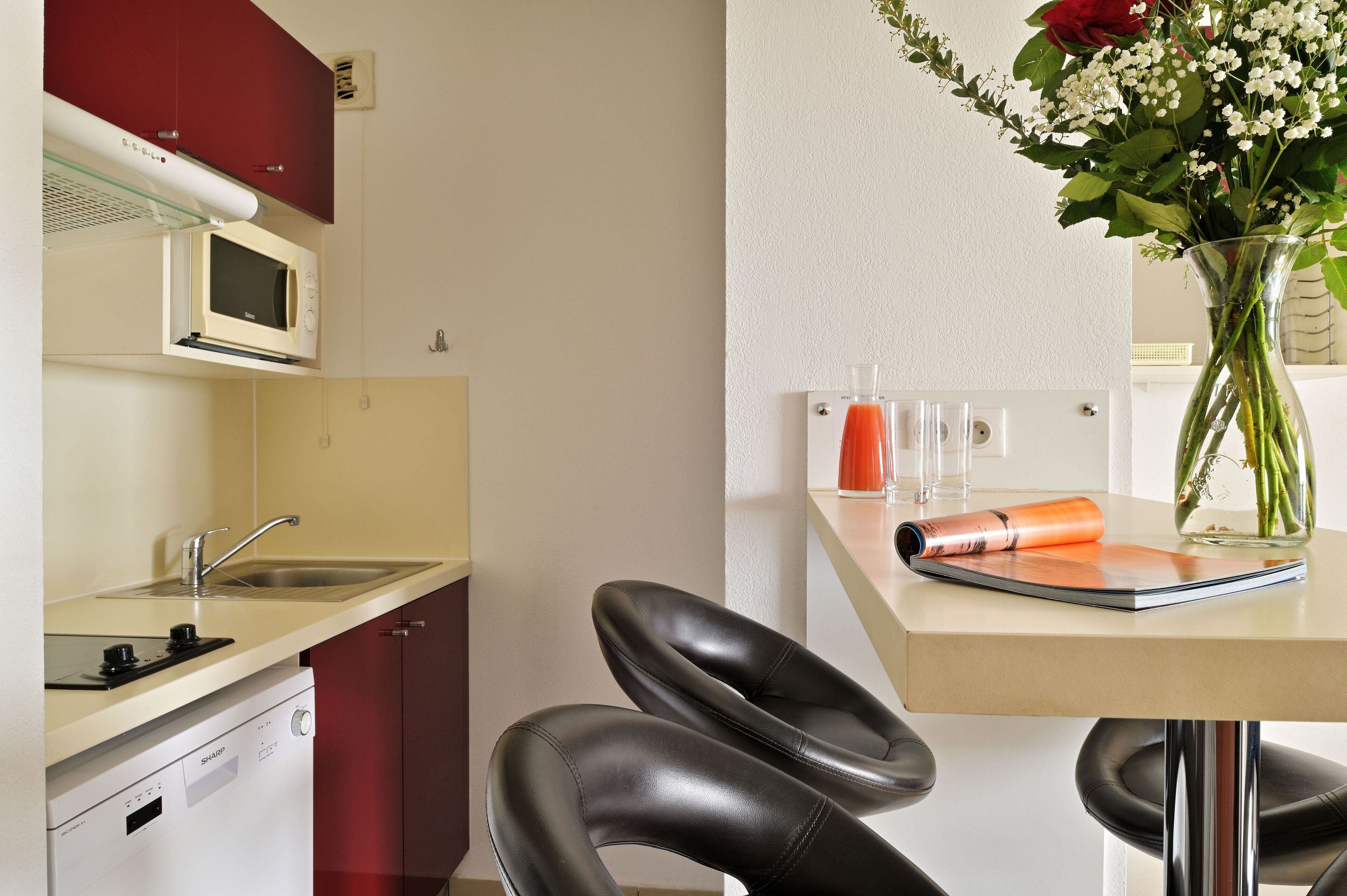A compact kitchen with a microwave, sink, and dishwasher. There's a dining table with chairs and a vase with flowers.